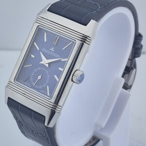 Pu&ograve; includere: Un orologio da polso rettangolare con quadrante blu e cassa color argento. L'orologio ha un cinturino in pelle blu scuro con motivo a coccodrillo. L'orologio &egrave; esposto su un supporto bianco.