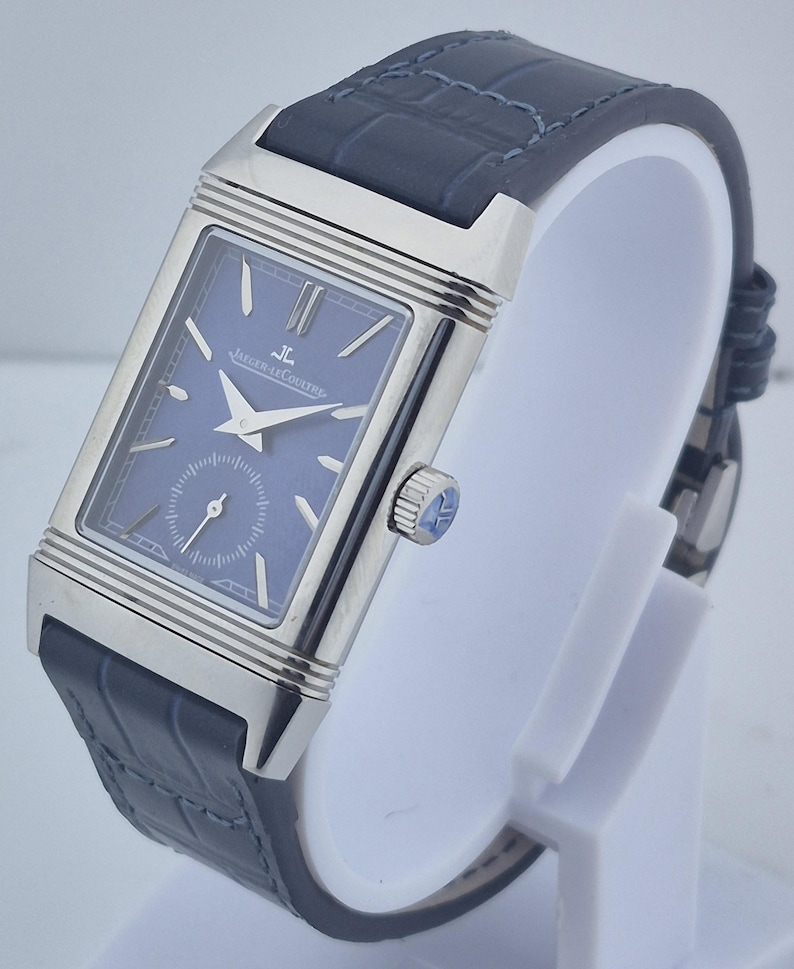 Pu&ograve; includere: Un orologio da polso rettangolare con quadrante blu e cassa color argento. L'orologio ha un cinturino in pelle blu scuro con motivo a coccodrillo. L'orologio &egrave; esposto su un supporto bianco.