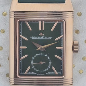 May include: A rectangular rose gold wristwatch with a dark green face and a small seconds dial. The watch features gold hands and markers, and a green leather strap. The watch face has the text "JAEGER-LECOULTRE" and "SWISS MADE".