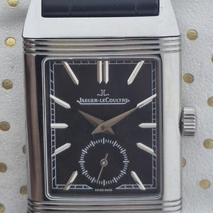Puede incluir: Reloj de pulsera rectangular con esfera negra y caja plateada. El reloj presenta la marca "JAEGER-LECOULTRE" y el texto "SWISS MADE". Tiene una correa de cuero negro.