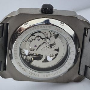 May include: Close-up of the back of a stainless steel automatic watch. The watch has a transparent window revealing the intricate inner workings. The watch is water resistant and features the brand name "BVLGARI".