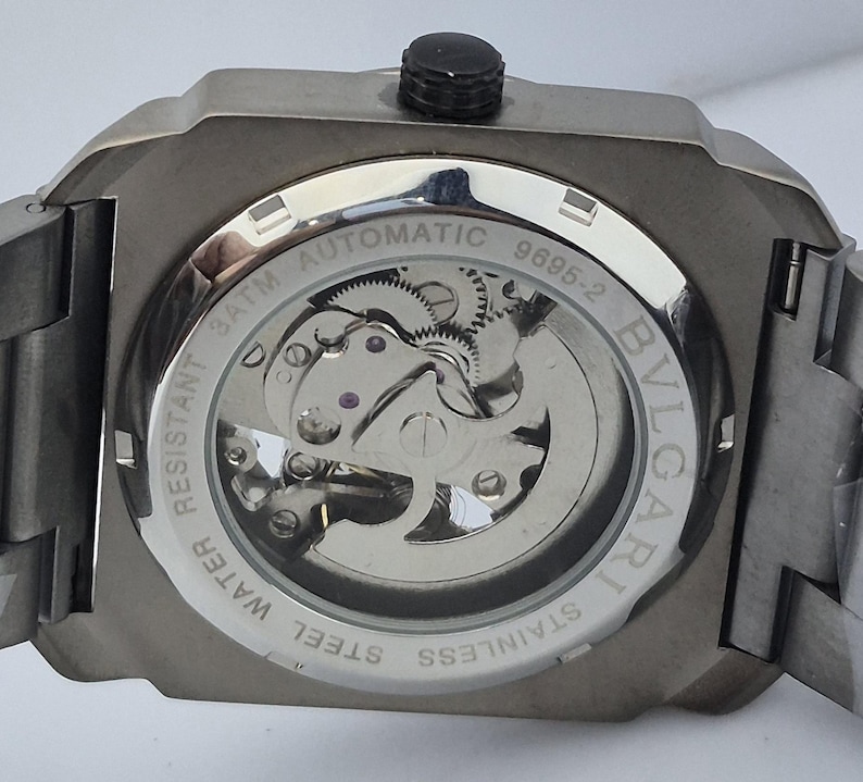 May include: Close-up of the back of a stainless steel automatic watch. The watch has a transparent window revealing the intricate inner workings. The watch is water resistant and features the brand name "BVLGARI".