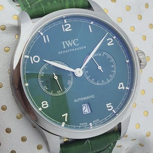 May include: A luxury wristwatch with a teal face and a green leather band. The watch features the brand name "IWC SCHAFFHAUSEN" and the word "AUTOMATIC". The watch has a date display and a second dial. The watch is a stylish accessory.