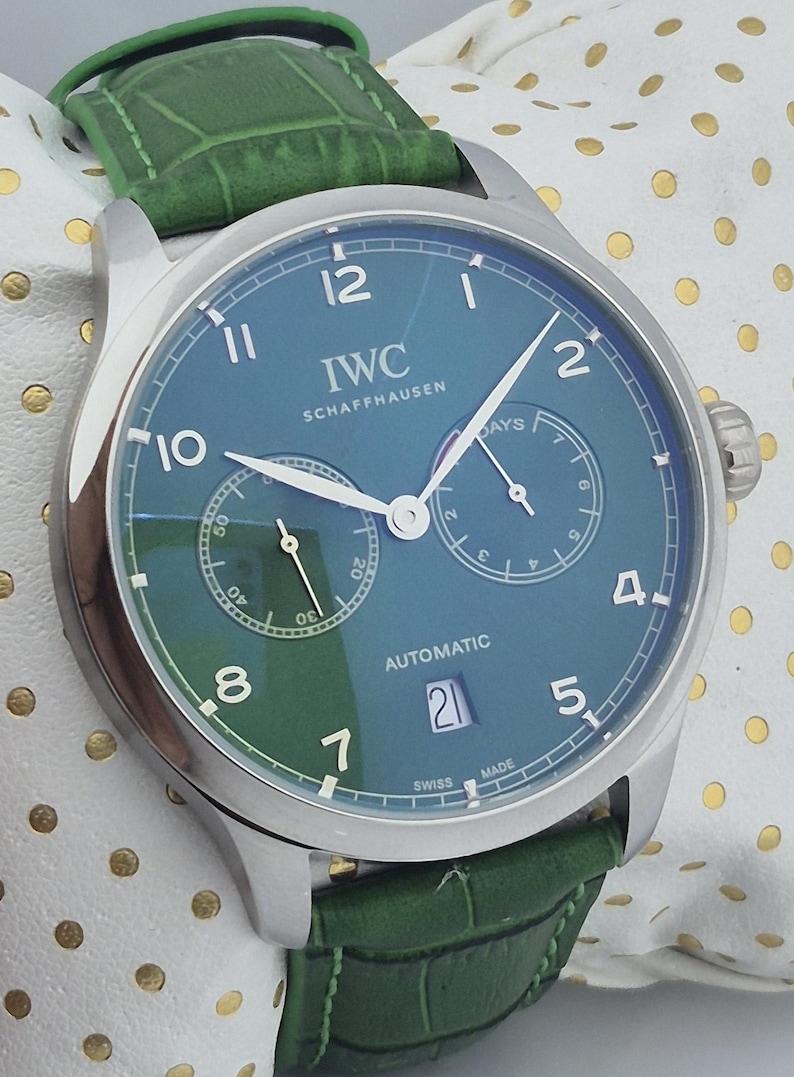 May include: A luxury wristwatch with a teal face and a green leather band. The watch features the brand name "IWC SCHAFFHAUSEN" and the word "AUTOMATIC". The watch has a date display and a second dial. The watch is a stylish accessory.