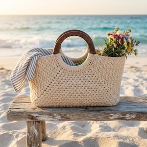 Puede incluir: Un bolso tote de macramé beige con asas de madera descansa sobre un banco de madera desgastada en la playa. La bolsa contiene una toalla a rayas y un ramo de flores coloridas. El océano es visible en el fondo.