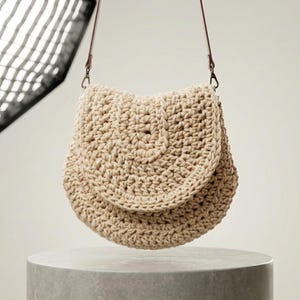 May include: A beige crochet bag with a flap closure and a brown leather strap. The bag is suspended in the air, showcasing its handmade texture and design. The bag is sitting on a gray cylindrical pedestal.