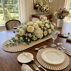 May include: A dining table set with a cream-colored oval crochet runner and matching round placemats. A large floral centerpiece features white and pale pink roses, hydrangeas, and greenery. White plates with gold rims, silverware, and crystal glasses.