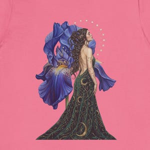 May include: A pink short-sleeve t-shirt featuring a graphic print of a figure in a long dark green dress with gold crescent moon patterns, standing in front of a large blue iris flower with a faint white dotted circle halo behind the head.