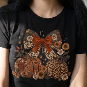 Könnte beinhalten: Ein schwarzes Kurzarm-T-Shirt mit einem Grafikdruck einer großen Schleife mit Leopardenmuster und Blumenmuster, zentriert über zwei Kürbissen. Ein Kürbis hat ein Blumenmuster, der andere ein Leopardenmuster, umgeben von Sonnenblumen.