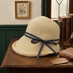 May include: A beige straw hat with a rounded crown and a slightly downturned brim. The hat features a dark navy blue ribbon band wrapped around the base of the crown, tied into a decorative bow on the side with two long, trailing ribbon ends.