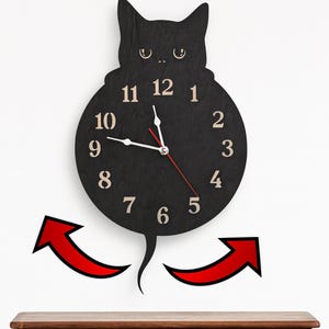 May include: A black cat-shaped wall clock with a round clock face. The clock features a black body with beige numbers and hands. The second hand is red. The clock is mounted on a white wall above a wooden surface.