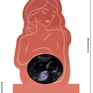 May include: A coral-coloured wooden frame in the shape of a pregnant woman, 192.5 mm wide and 245 mm tall, with a sonogram image in the belly. The base is 83.5 mm wide. The frame features a minimalist design.