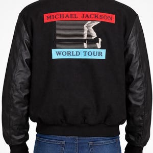 Puede incluir: Chaqueta bomber negra con mangas de cuero. La parte trasera presenta un gráfico de la silueta de Michael Jackson y las palabras "MICHAEL JACKSON WORLD TOUR" en rojo y azul.