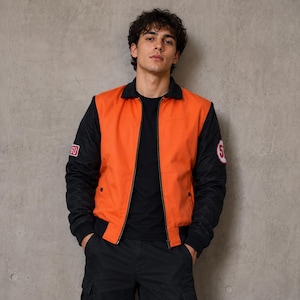 Puede incluir: Chaqueta bomber naranja y negra con cuello y puños negros. La chaqueta tiene parches en las mangas. La persona de la imagen lleva una camiseta negra y pantalones negros.