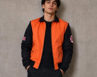 Könnte beinhalten: Orange-schwarze Bomberjacke mit schwarzem Kragen und Manschetten. Die Jacke hat Aufnäher auf den Ärmeln. Die Person im Bild trägt ein schwarzes T-Shirt und eine schwarze Hose.