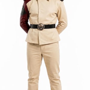May include: A beige jacket with a collar, dark red right sleeve, and black shoulder epaulets, paired with matching beige trousers. A black belt with a circular silver skull buckle is worn at the waist. The outfit includes beige lace-up boots.