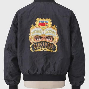 May include: Black bomber jacket featuring a colorful graphic on the back. The graphic includes the Pepsi logo, the words "Michael Jackson" and "Dangerous World Tour" in gold lettering, and an image of Michael Jackson's eyes.