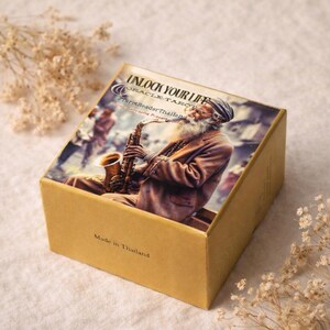 May include: A gold-colored box featuring an image of a man with a long white beard playing a saxophone. The box has the text "UNLOCK YOUR LIFE" and "Made in Thailand". The box is decorated with dried flowers.