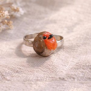 Puede incluir: Anillo de plata con un petirrojo pintado a mano. El pájaro tiene un cuerpo marrón, un pecho naranja brillante y ojos negros. El anillo está sobre un fondo texturizado blanquecino.
