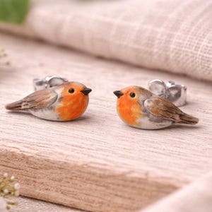 Könnte beinhalten: Zwei handgefertigte Rotkehlchen-Figuren mit orangefarbenen Brüsten, braunen Flügeln und schwarzen Schnäbeln. Die Vögel sitzen auf einer hellen Holzoberfläche, an der jeweils ein silberner Ring befestigt ist. Im Hintergrund weiße Blüten.