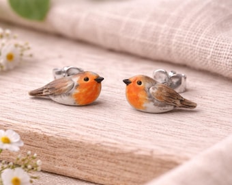 May include: Two handcrafted robin bird figurines with orange breasts, brown wings, and black beaks. The birds are perched on a light-colored wooden surface, with a silver ring attached to each bird. The background includes white flowers.