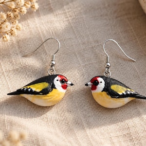 Op de afbeelding: Een paar hangende oorbellen met miniatuur putter-bedels. Elke vogel is beschilderd met een zwarte kop, een felrode vlek op het gezicht, een witte nek en gele en zwarte vleugels. De bedels hangen aan zilverkleurige metalen oorhaken.