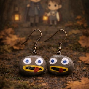 Puede incluir: Pendientes hechos a mano con un diseño caprichoso. Cada pendiente es una piedra gris y redondeada con una cara pintada: ojos azules, boca amarilla y lengua roja. Los pendientes cuelgan de ganchos dorados.