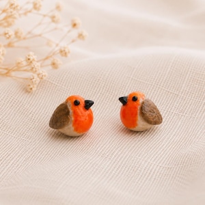 Puede incluir: Dos figuras en miniatura de petirrojos con pechos de color naranja brillante, alas marrones y pequeños picos negros. Cada ave tiene un cuerpo liso y redondeado con un acabado brillante, detallado con diminutos ojos negros, lado a lado.
