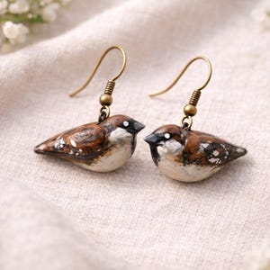 Puede incluir: Un par de pendientes artesanales con forma de pájaro. Cada pendiente presenta un pequeño y detallado gorrión con colores marrón, blanco y negro. Los pendientes tienen ganchos dorados. El fondo es una tela suave y neutra.