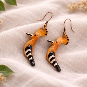 Puede incluir: Un par de pendientes hechos a mano con diseño de pájaro. Los pendientes son de color naranja, negro y blanco, con ganchos para las orejas de color cobre. La cabeza del pájaro es naranja con lunares negros, y el cuerpo tiene rayas blancas y negras.