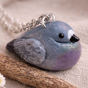 Op de afbeelding: Een handgemaakte vogelhangerketting. De hanger is een gedetailleerde sculptuur van een duif, met een blauwgrijs lichaam, iriserende accenten en een zwarte snavel. De vogel zit op een houten tak, met een zilveren ketting.