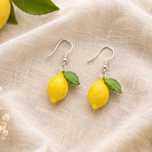 以下が含まれることがあります： 小さな緑の葉が付いたミニチュアの黄色いレモンのチャームが特徴のドロップイヤリング。レモンは銀色の金属製フックから吊り下げられています。このデザインは、果物の質感のある表面と葉の鮮やかな緑色を捉えており、コンパクトで身につけやすい形でレモンをリアルに表現しています。