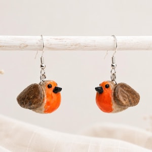 Puede incluir: Un par de pendientes hechos a mano con pequeñas figuras detalladas de petirrojos. Cada ave tiene un pecho de color naranja brillante, alas marrones y un pico negro. Los pendientes cuelgan de ganchos plateados, mostrando un diseño inspirado en la naturaleza.