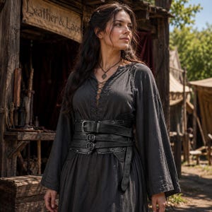 Op de afbeelding: Een donkergrijze tuniek met lange mouwen, een vetersluiting bij de hals en een brede zwarte leren korsetgordel met meerdere riemen, zilverkleurige gespen en metalen studs, gedragen over de tuniek om de taille strak aan te snoeren.