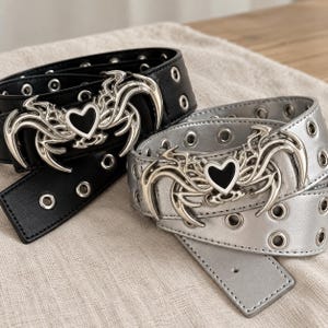 May include: Two belts, one black and one silver, featuring rows of metal grommets along the length. Each belt has a large, intricate silver-toned buckle shaped like a stylised spider with a central black heart-shaped accent at its core.
