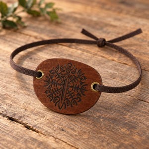 May include: A brown leather eye patch featuring a dark engraved design of a sword centered within a circular geometric pattern. The patch is attached to a thin, dark brown fabric cord that threads through two brass-colored metal eyelets.