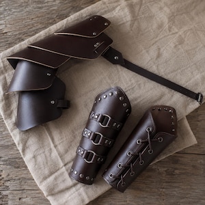 May include: A set of dark brown leather armour pieces including a multi-layered shoulder pauldron with a long adjustable strap and two forearm bracers. One bracer features three metal ring buckles, while the other has a crisscross lace-up closure.