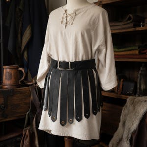 May include: A beige long-sleeved tunic with a lace-up neckline paired with a black leather belt featuring long, vertical hanging strips and metallic studs at the bottom of each strip, secured with a silver-toned metal buckle at the waist.