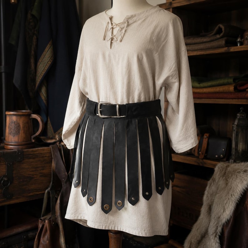 May include: A beige long-sleeved tunic with a lace-up neckline paired with a black leather belt featuring long, vertical hanging strips and metallic studs at the bottom of each strip, secured with a silver-toned metal buckle at the waist.