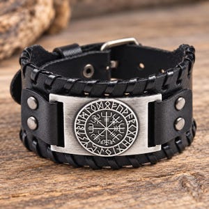 Puede incluir: Una pulsera ancha de cuero negro con una placa metálica rectangular de tono plateado en el centro. La placa muestra un símbolo circular Vegvisir rodeado de caracteres rúnicos. La banda de cuero tiene bordes cosidos y un cierre de hebilla.