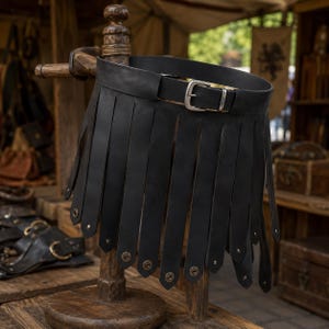 May include: A black leather belt featuring a wide waistband with a silver-toned metal buckle and a series of long, vertical leather strips hanging from the bottom edge, each finished with a small, circular metallic stud at the pointed tip.
