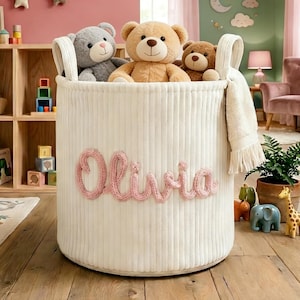 May include: A cream-colored, corduroy storage basket with the name "Olivia" in pink script. The basket is filled with three teddy bears. A wooden shelf with books and a teddy bear is in the background.