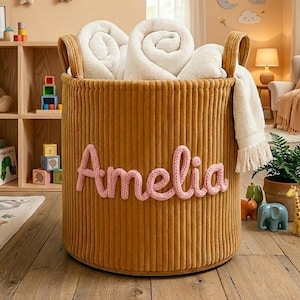 May include: A round, mustard-colored corduroy storage basket with the name "Amelia" in pink script. The basket is filled with white blankets and has two handles. A children's room setting with a wooden bookshelf, toys, and a rug.