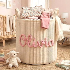 Puede incluir: Una cesta de almacenamiento de pana color crema con el nombre "Olivia" en rosa. La cesta está llena de ropa doblada. Una alfombra redonda de yute, un juguete de madera y un cordero de peluche están en primer plano. Una cuna y arte enmarcado están en el fondo.