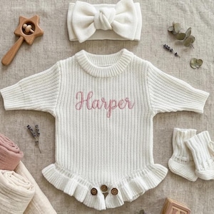 Op de afbeelding: Een witte gebreide babyromper met de naam "Harper" in roze geborduurd, een bijpassende hoofdband en sokken. De romper heeft een gefranjerde zoom en houten knopen. Een houten ster-vormige rammelaar en een blok zijn ook aanwezig.