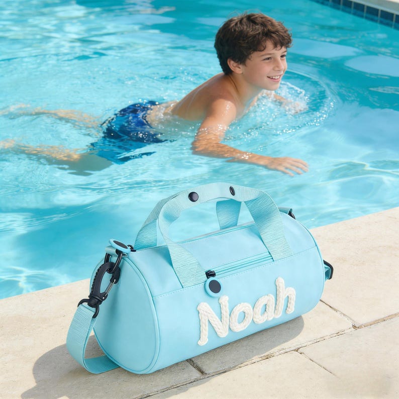 May include: A pale blue duffel bag with the name "Noah" embroidered in white rope-like letters. The bag has a top handle and a detachable shoulder strap. A young boy is swimming in a pool in the background.