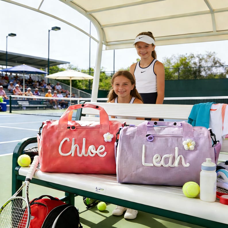 May include: Two personalised duffel bags, one coral and one lavender, sit on a bench at a tennis court. The coral bag has "Chloe" embroidered, and the lavender bag has "Leah". Both bags have a flower accent.