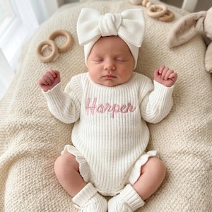 Puede incluir: Un bebé recién nacido con un pelele de punto blanco con el nombre "Harper" bordado en rosa. El conjunto incluye una diadema con lazo y calcetines a juego. Juguetes de madera y un peluche en el fondo.