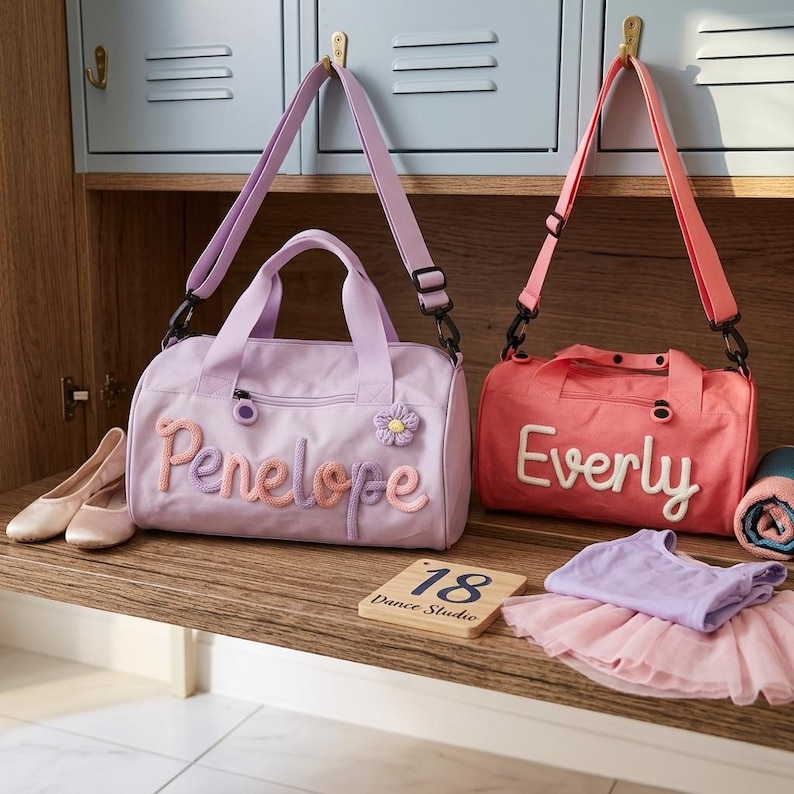 May include: Two small duffel bags, one lilac and one coral, with adjustable shoulder straps. The lilac bag has the name "Penelope" embroidered on it, and the coral bag has the name "Everly". Ballet shoes, a tutu, and a wooden sign that says "18 Dance Studio" are also present.