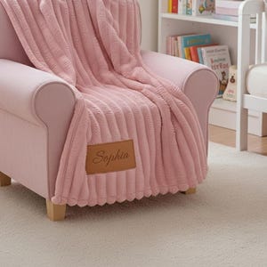 May include: A pink, ribbed throw blanket draped over a pink armchair. The blanket has a leather patch with the name "Sophia" stitched on it. The chair is set on a light-colored carpet, with a bookshelf and a crib in the background.